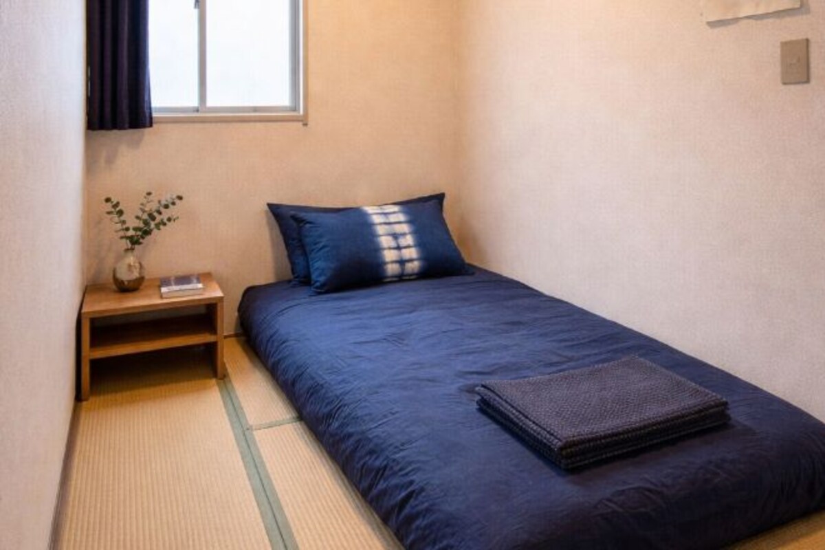 A small, neatly arranged bedroom with a single bed, a nightstand, a small table, and a window with curtains.