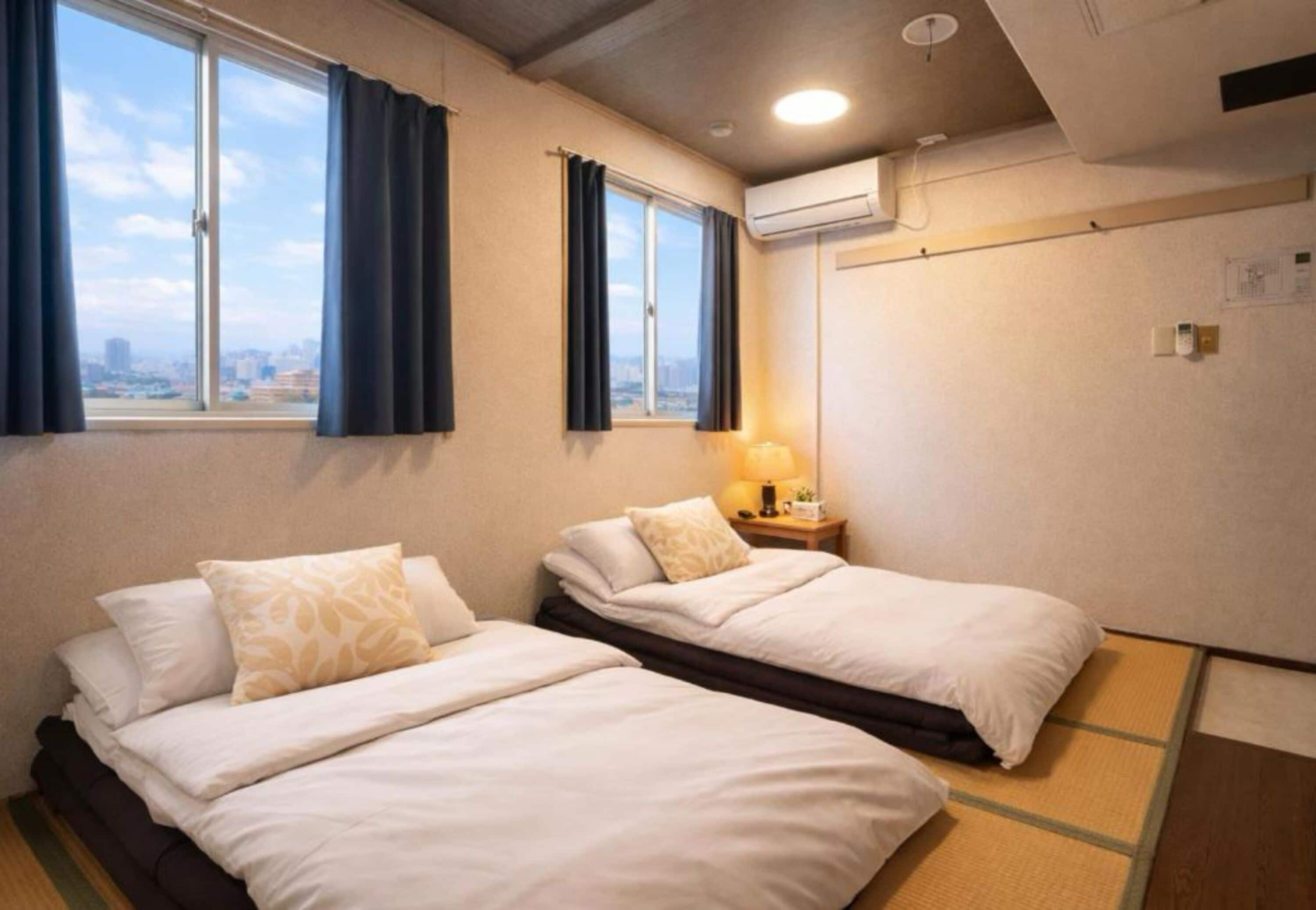A hotel room with two beds, a window with curtains, an air conditioning unit, and a bedside table with a lamp.