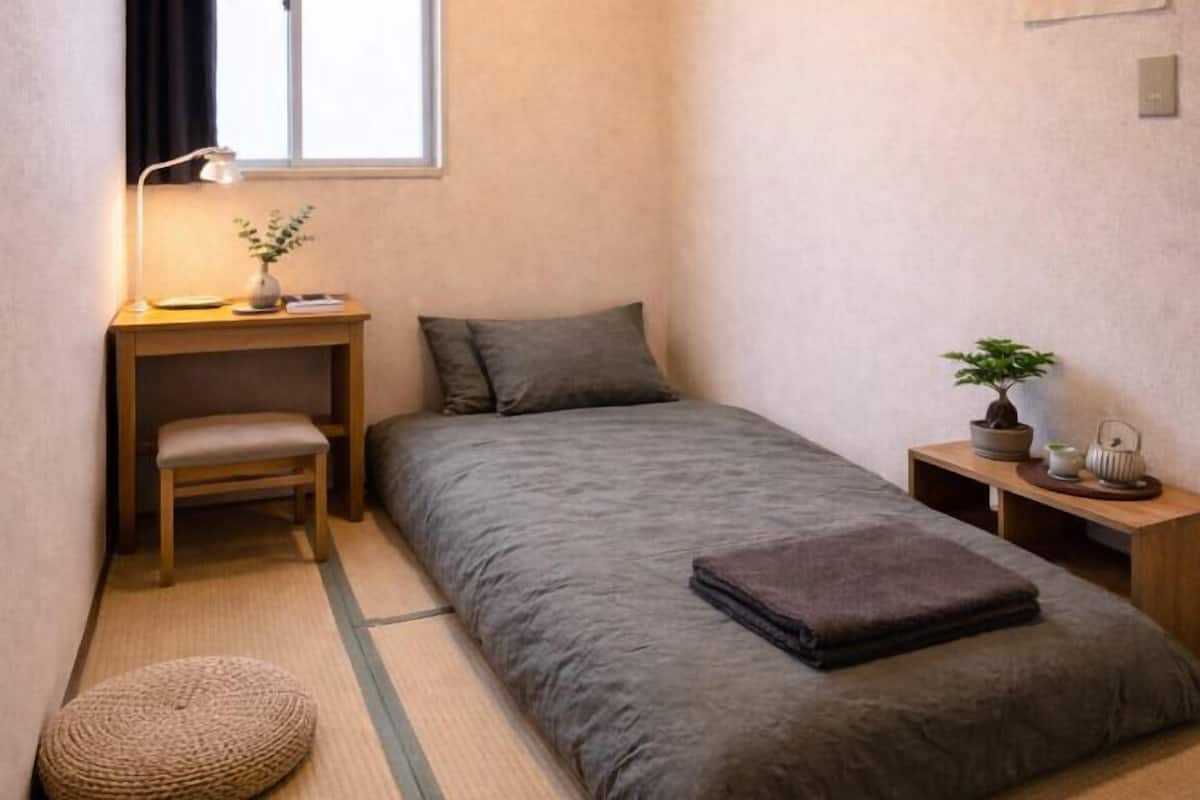 A small, neatly arranged bedroom with a bed, a chair, a desk with a lamp, and a small table with a plant.