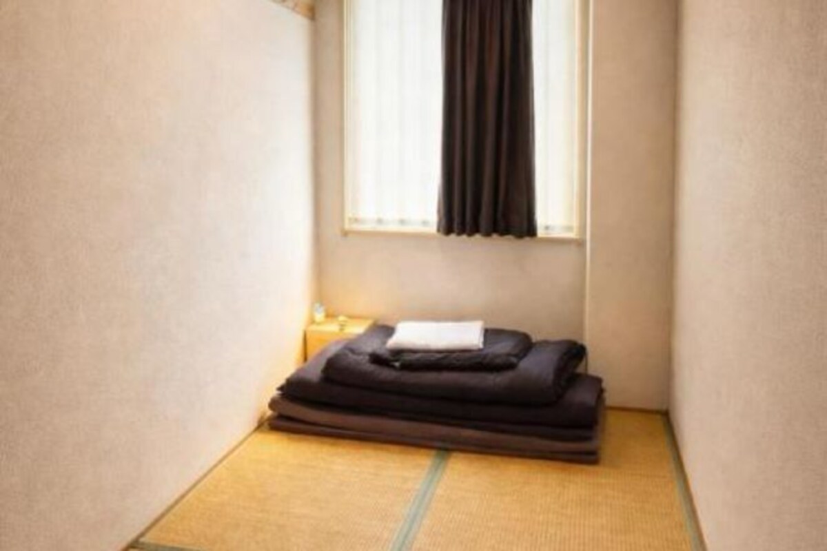 A small room with a tatami mat floor, a window with curtains, and a stack of folded blankets.