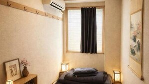 Una habitación japonesa tradicional con suelo de tatami, un futón, una pequeña mesa de madera con un jarrón y un aire acondicionado de pared.