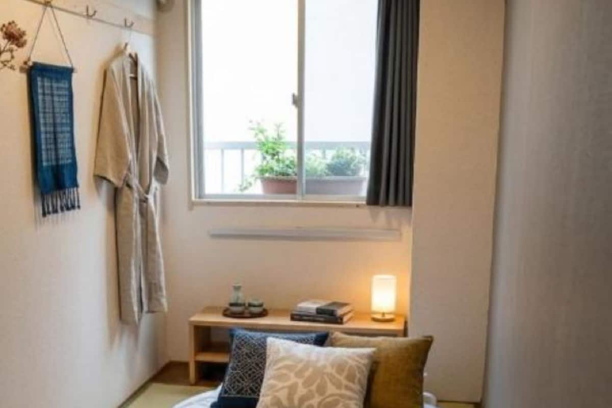 A small, cozy bedroom with a single bed, a bedside table with a lamp, and a window with a view of the outside.