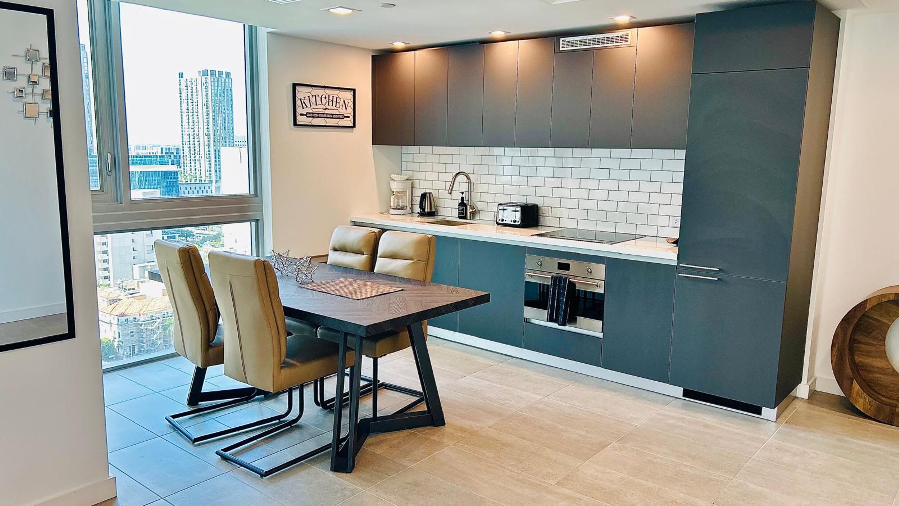 A modern kitchen with a dining area, featuring a kitchen island, dark cabinets, and a city view through the window.