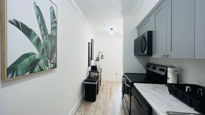 Un couloir étroit, avec un coin cuisine sur la droite, comprenant un four à micro-ondes, une cuisinière et des placards. On peut apercevoir une poubelle et une petite table avec une chaise dans le couloir.