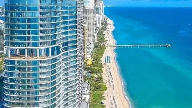 Um hotel com fachada curva e muitos andares, situado ao lado de uma praia e um cais.