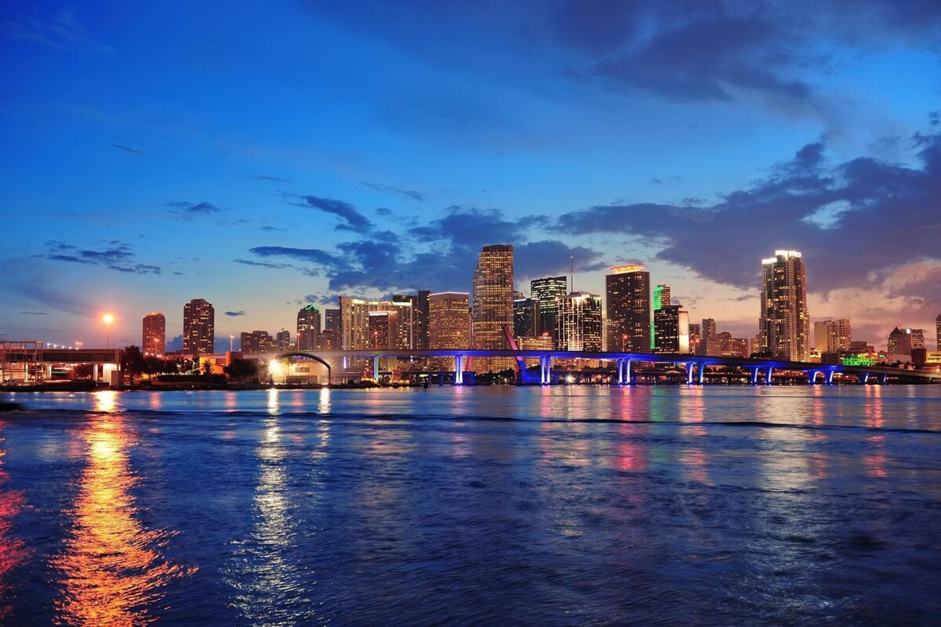 A city skyline at dusk with illuminated buildings and a bridge over a body of water.