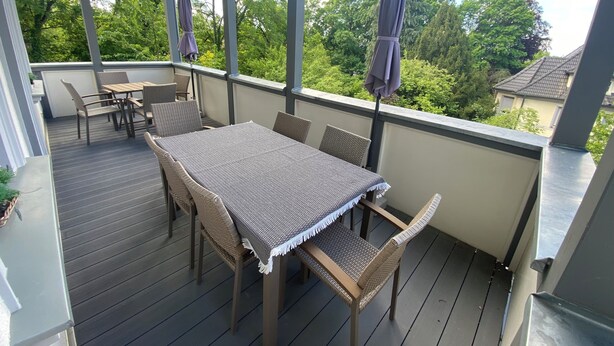 A covered outdoor patio with a table and chairs, surrounded by large windows offering a view of trees and a house.