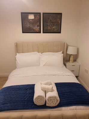 A neatly made bed with white linens and a blue blanket, a beige headboard, and two framed floral artworks on the wall.