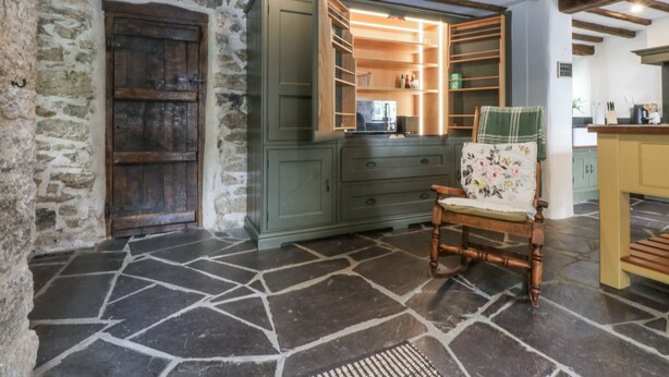 A rustic room with stone flooring, wooden beams, a green cabinet, and a chair with a floral cushion.