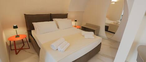 A neatly made bed with white linens and rolled towels, a small orange table with a lamp, and a mirror in the corner of the room.