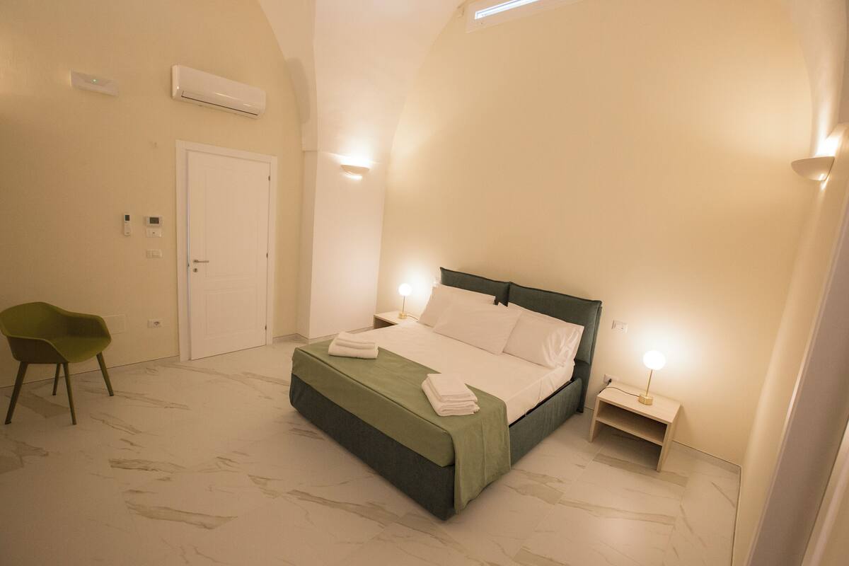 A bedroom with a bed, bedside tables, a chair, and an air conditioning unit.