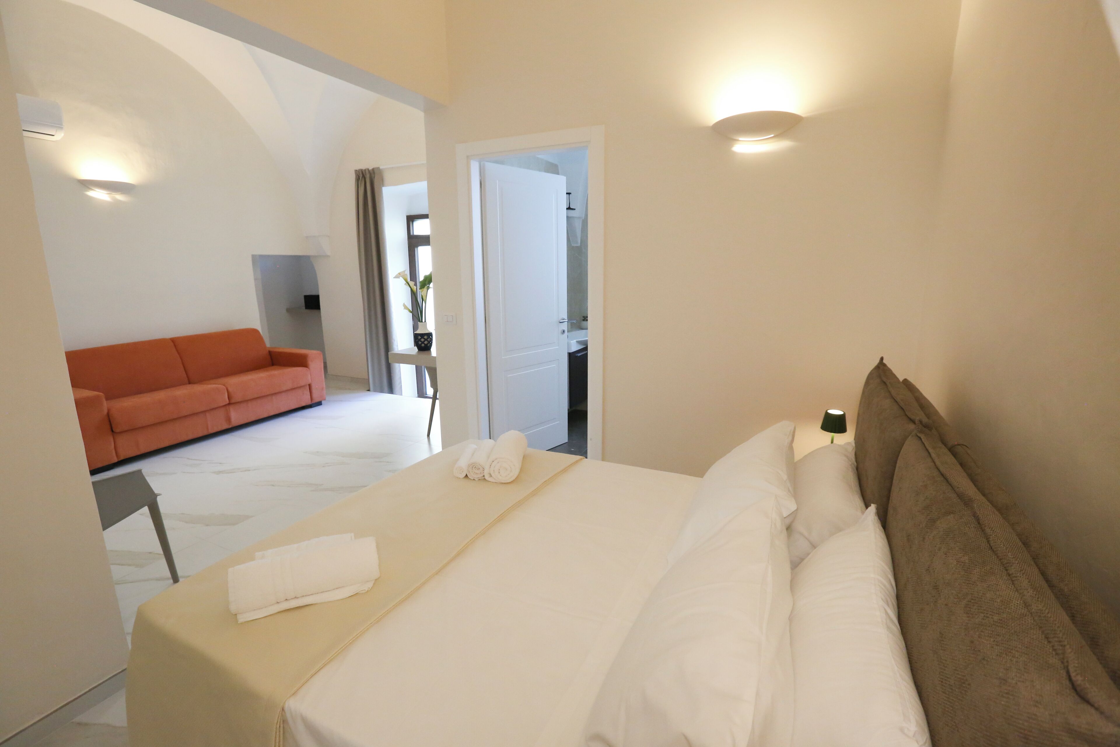 Habitación de hotel con cama, sofá y baño visible a través de una puerta abierta.