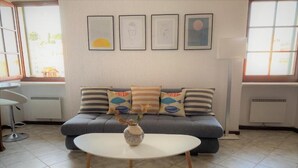 A living room with a gray sofa, a white coffee table, and a vase with dried flowers. There are framed pictures on the wall and a window with blinds.