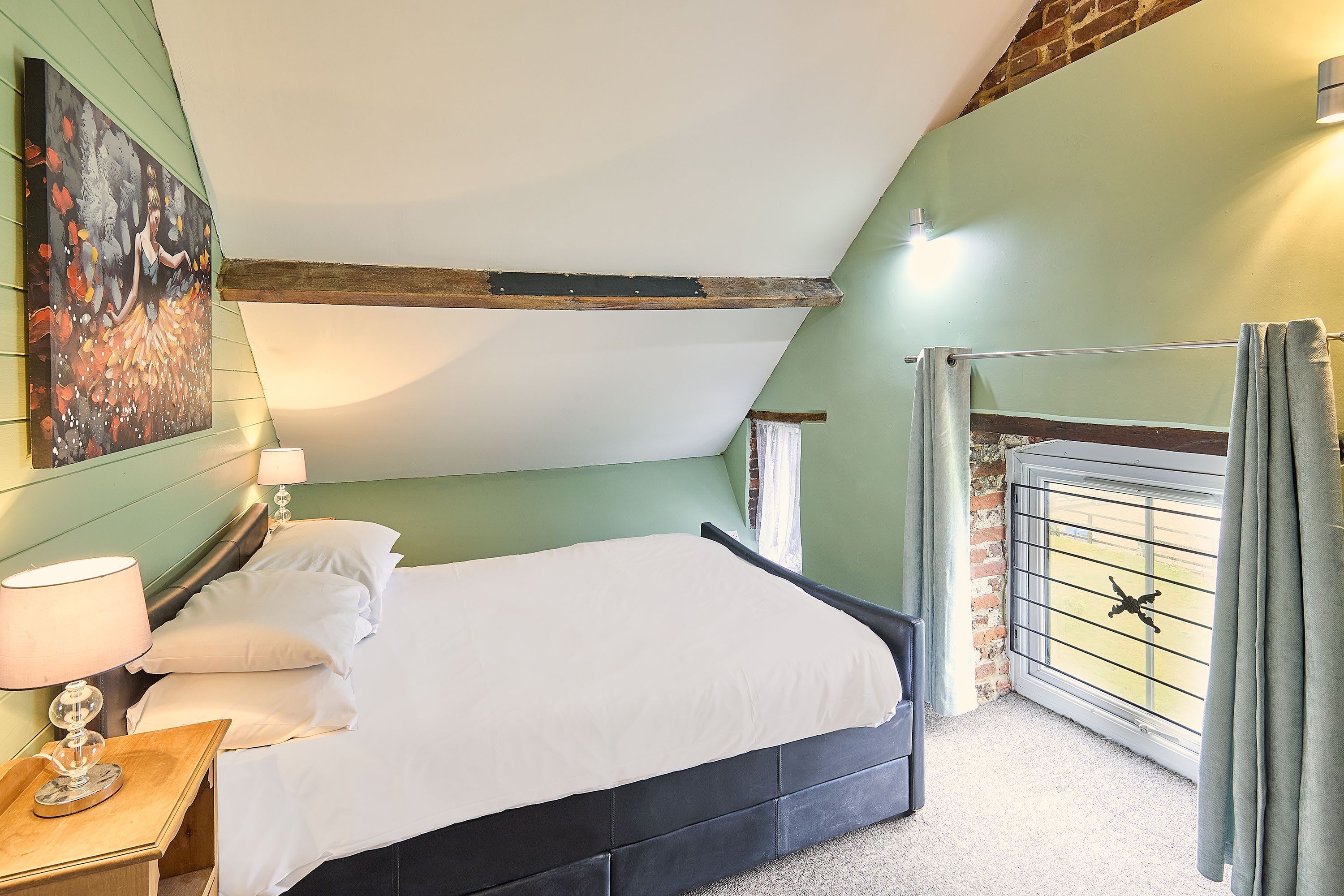 Een slaapkamer met een bed, nachtkastje, schilderij en raam met gordijnen.