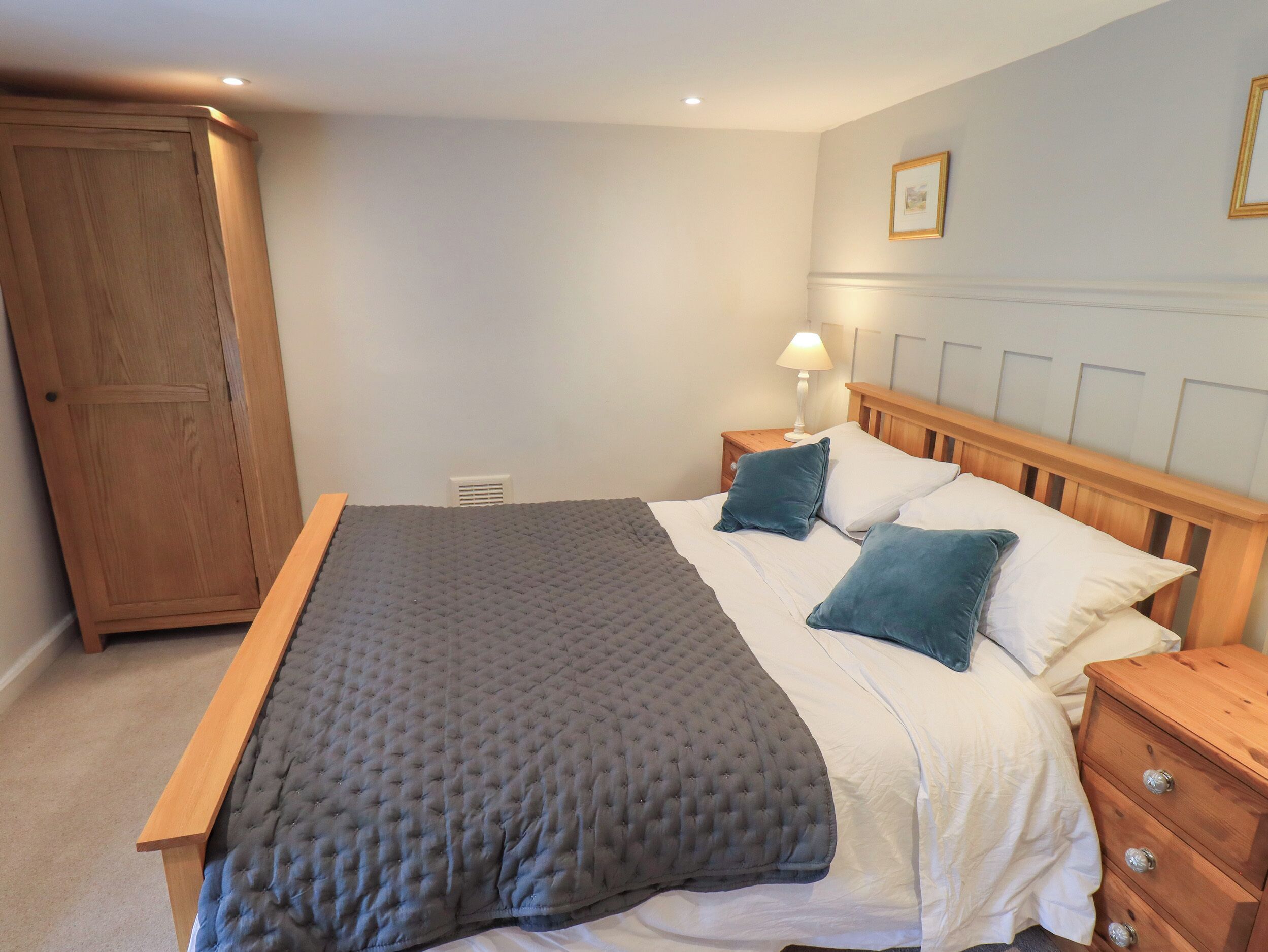 A bedroom with a large bed, wooden headboard, a nightstand with a lamp, a wardrobe, and a wall-mounted picture frame.