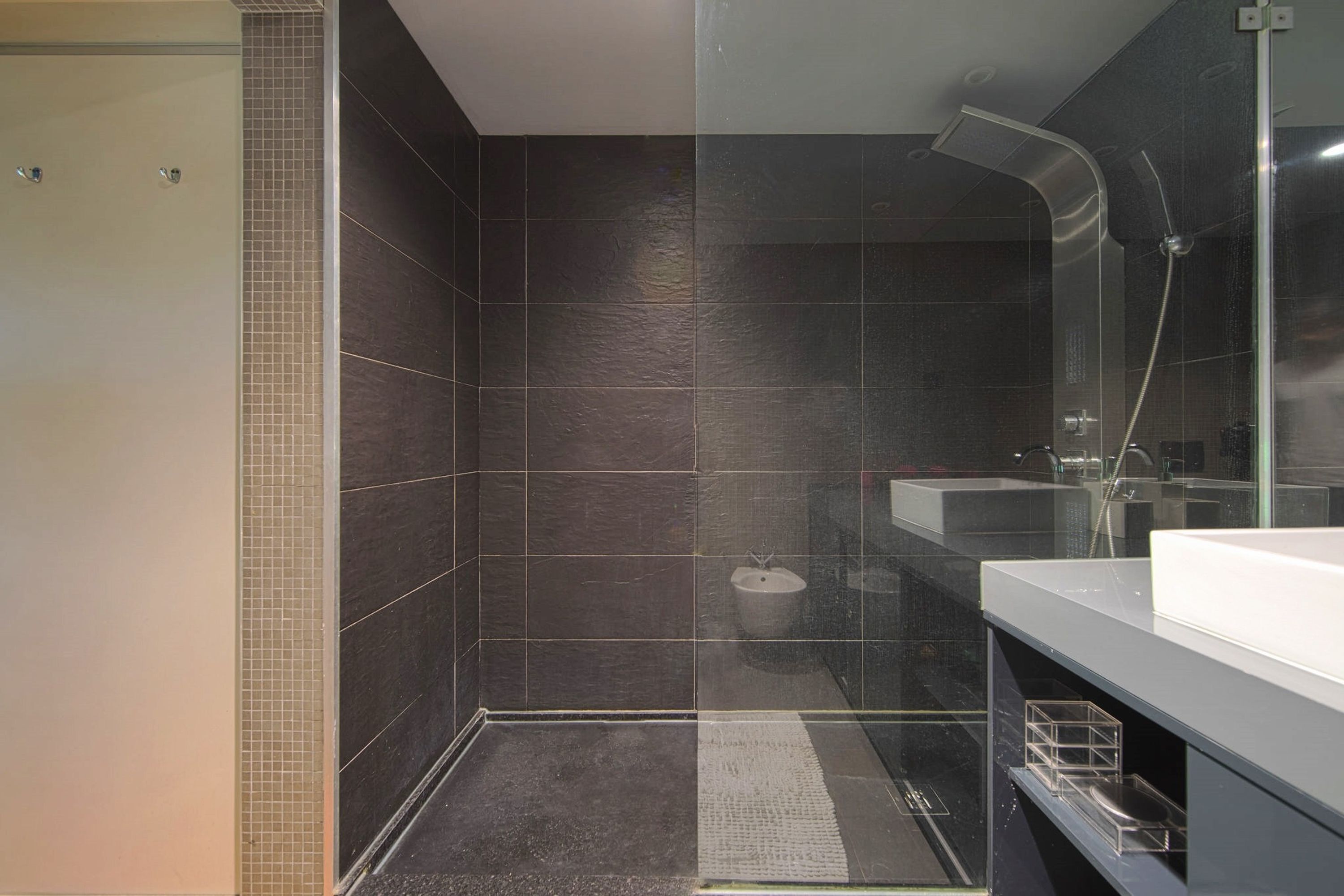 Ein modernes Badezimmer mit dunklen Fliesen, einer Glasdusche, einem Waschbecken und einem Spiegel.