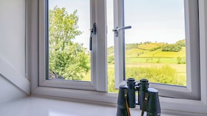 Ein Fenster mit Fernglas auf einer weißen Fensterbank, mit Blick auf eine grüne Landschaft mit Hügeln.