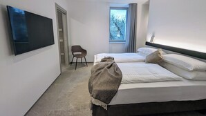 Une chambre moderne avec un grand lit, un téléviseur fixé au mur, un fauteuil et une fenêtre donnant sur la forêt.