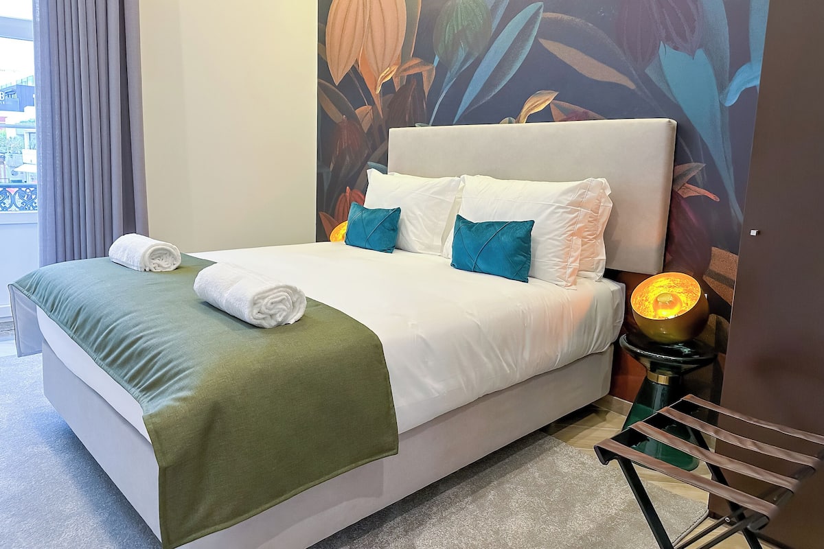 A neatly made bed with white linens and a green throw, a nightstand with a lamp, and a wall mural of tropical foliage.