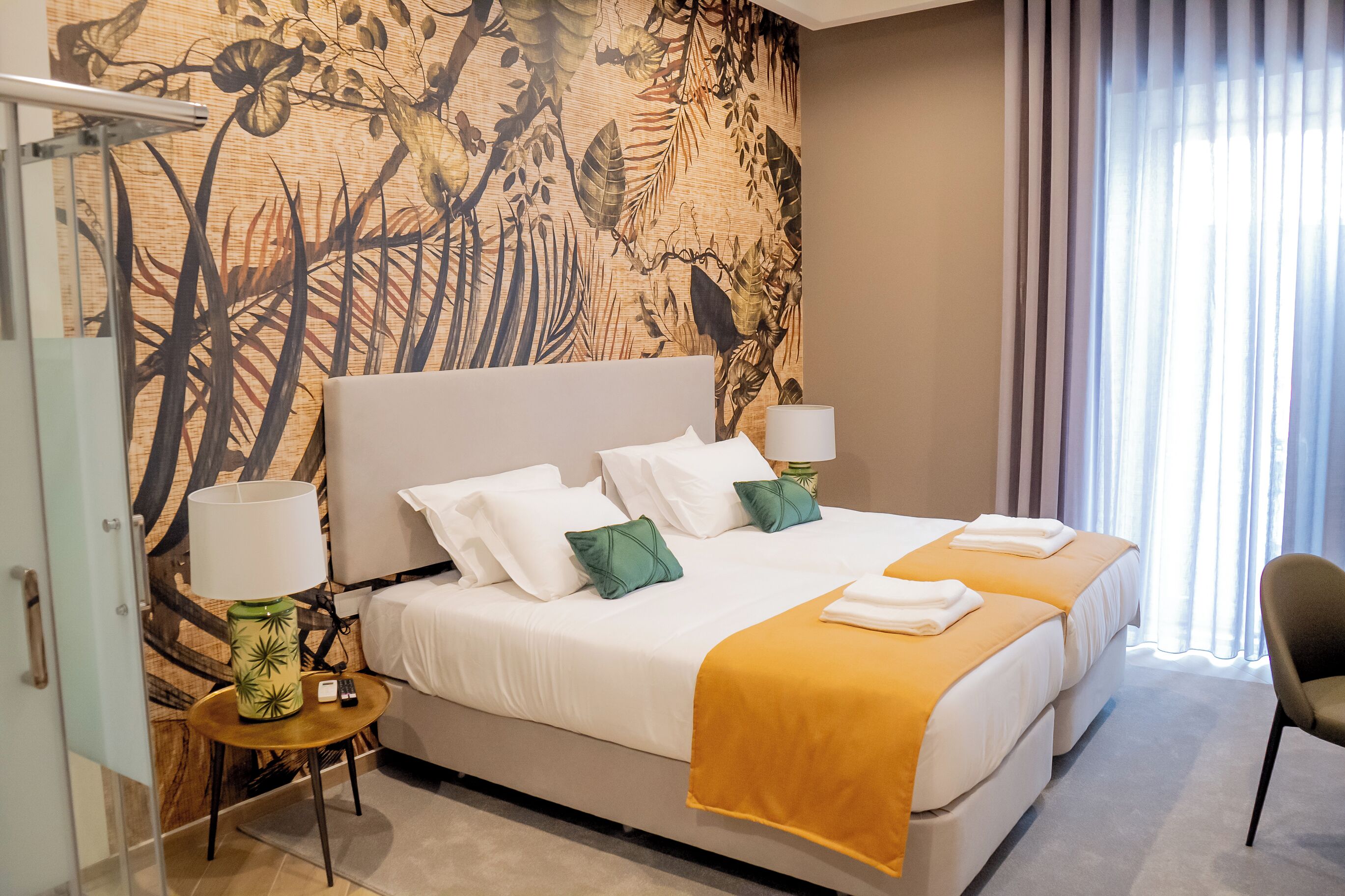 A hotel room with a large bed, two bedside tables, a lamp, and a view of the outdoors through sheer curtains.