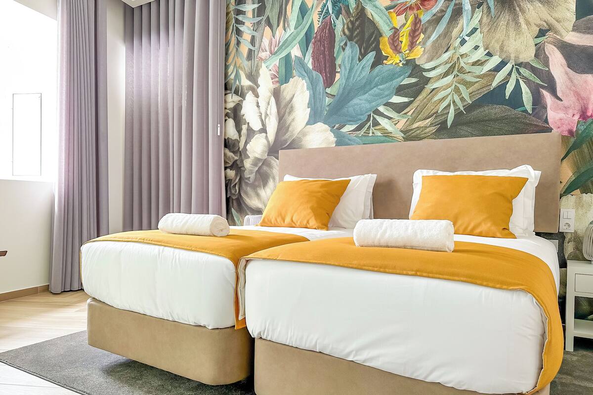A double bed with white and yellow bedding, a headboard with a floral mural, and a grey curtain.