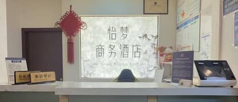 A reception desk with a sign that reads 'Yingma Business Hotel.' There are framed certificates on the wall, a red decorative hanging, and a computer monitor on the desk.