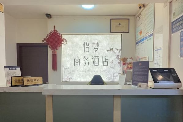 Een receptiebalie met een bordje met de tekst 'Yingma Business Hotel'. Er hangen ingelijste certificaten aan de muur, een rode decoratieve versiering en een beeldscherm op het bureau.