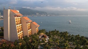 A coastal hotel with a view of the sea, a sailboat, and a plane in the distance.