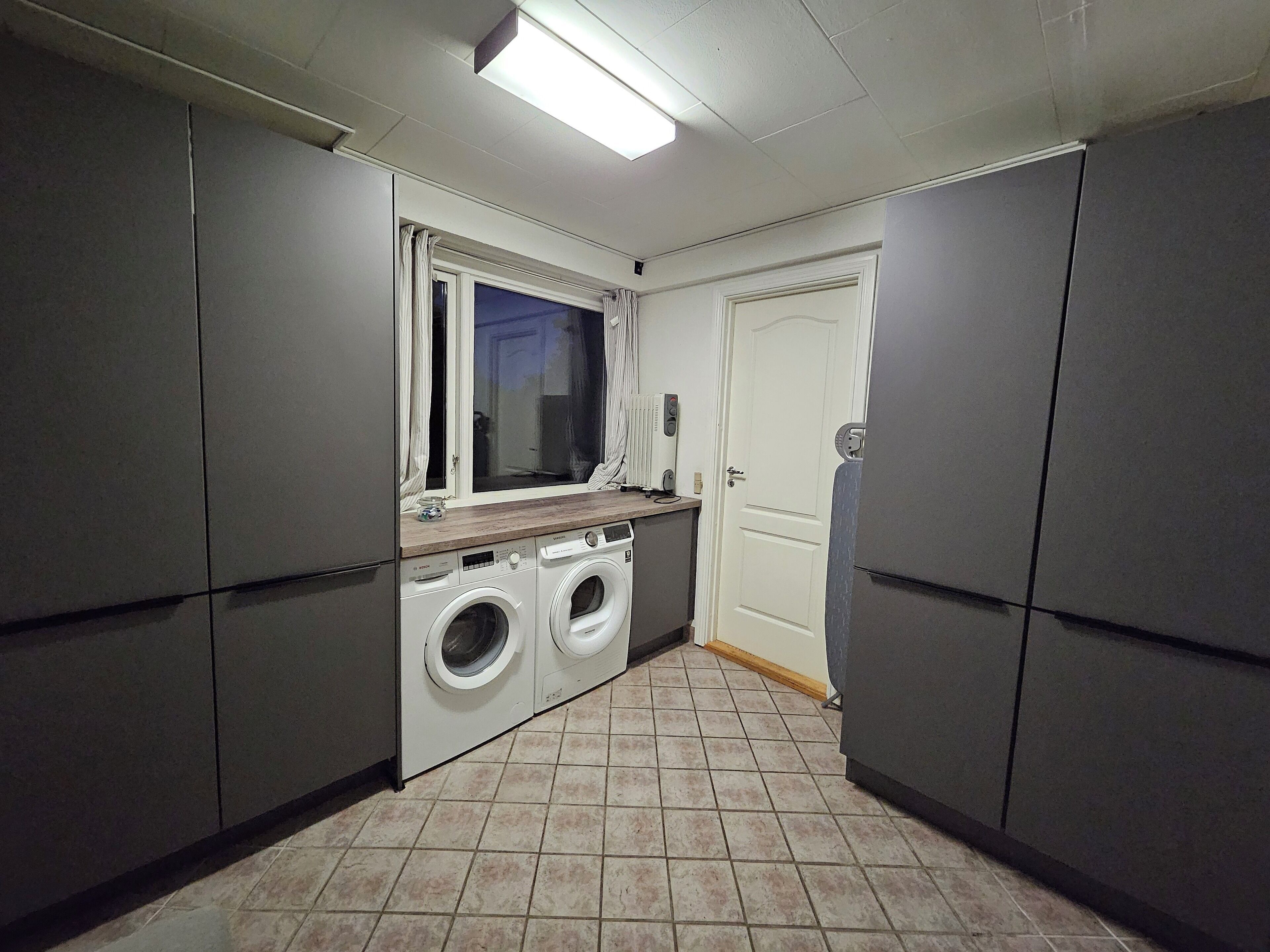 Une petite buanderie équipée de deux machines à laver, d’une fenêtre avec des rideaux et d’une porte.