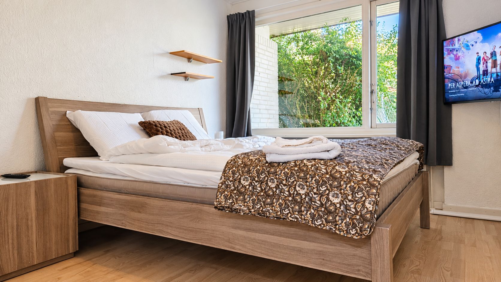 Un lit bien fait, recouvert d’une couverture à motifs, une table de chevet, un téléviseur fixé au mur et une fenêtre avec des rideaux.