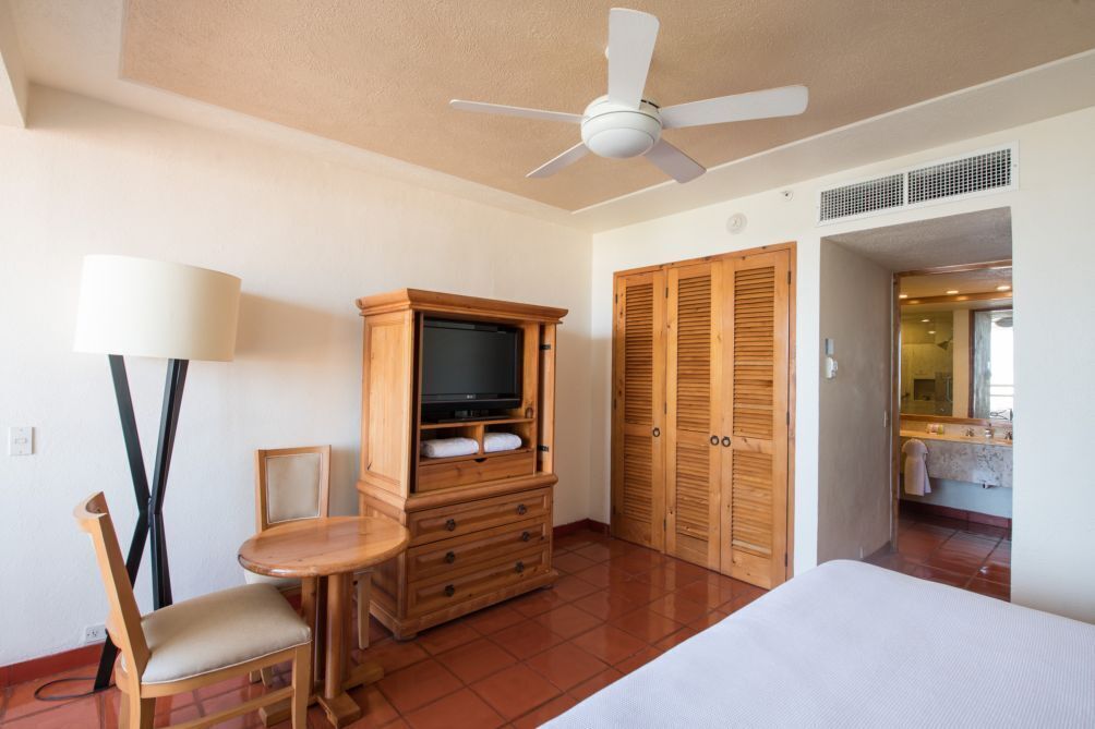 Habitación de hotel con cama, un mueble bajo de madera para la televisión con un televisor de pantalla plana, una pequeña mesa de madera con una silla, un ventilador de techo y armarios de madera.