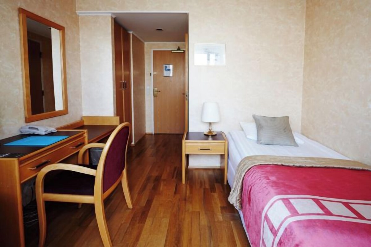 A hotel room with a bed, a desk with a chair, a bedside table with a lamp, and a door leading to another room.