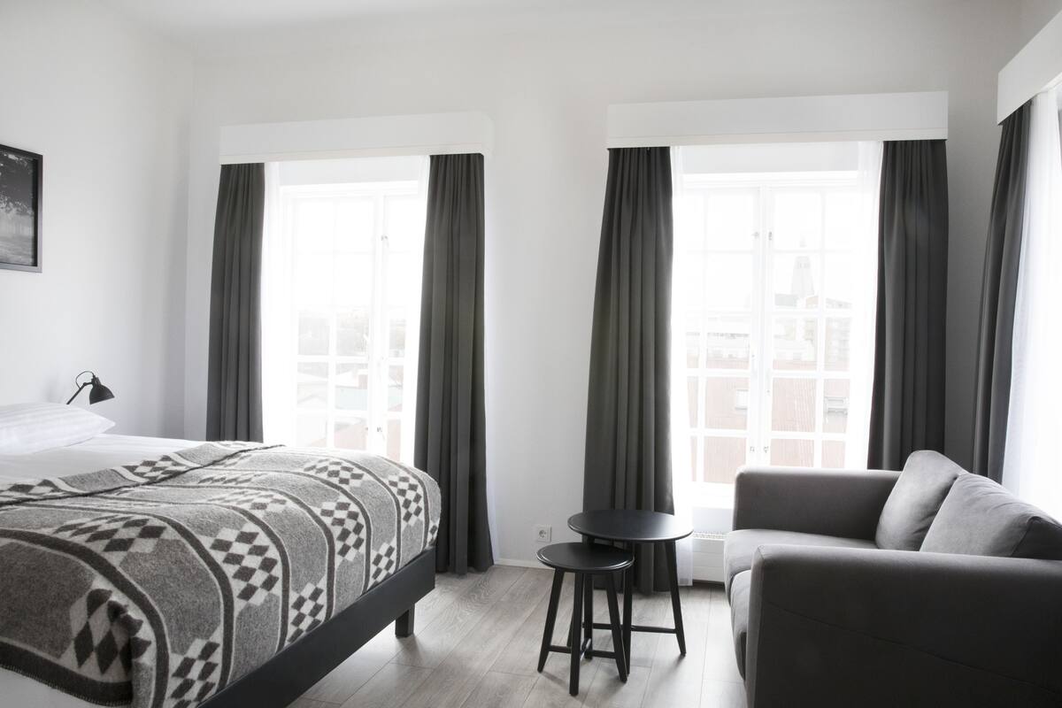 Una camera d'albergo moderna con un letto, una poltrona, un tavolino e ampie finestre con tende.