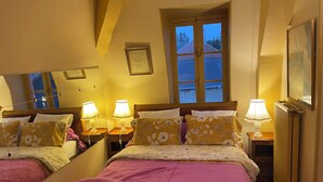 Una cama doble con ropa de cama floral, dos lámparas de noche, una ventana con vistas a edificios y un espejo en la pared.
