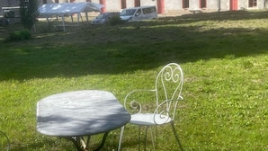 Un espacio al aire libre con césped, una mesa y silla de metal, un edificio con contraventanas rojas al fondo y un cielo despejado.