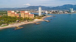 Un hotel frente al mar con varias construcciones, una playa y montañas al fondo.