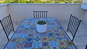 Une table ornée d’un motif de carreaux colorés, deux chaises et une plante en pot sur un balcon offrant une vue sur la ville.