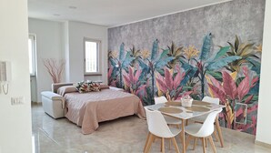 Une chambre à coucher moderne avec un lit, une table à manger avec des chaises, une grande fresque murale aux motifs tropicaux et une fenêtre.