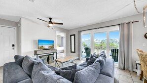 A modern living room with a grey sofa, a coffee table, and a TV. There is a balcony with a view of houses and greenery. - Beachside Villas 1121 (Santa Rosa Beach)