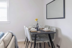A dining area with a table set for four, four chairs, a vase with a single flower, and a framed picture on the wall.
