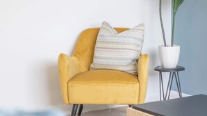 A yellow armchair with a striped pillow, a small round table with a potted plant, and a black table in a room with a light-colored carpet. - Leven Loft -  2 Bed Flat in Coatbridge (Coatbridge)