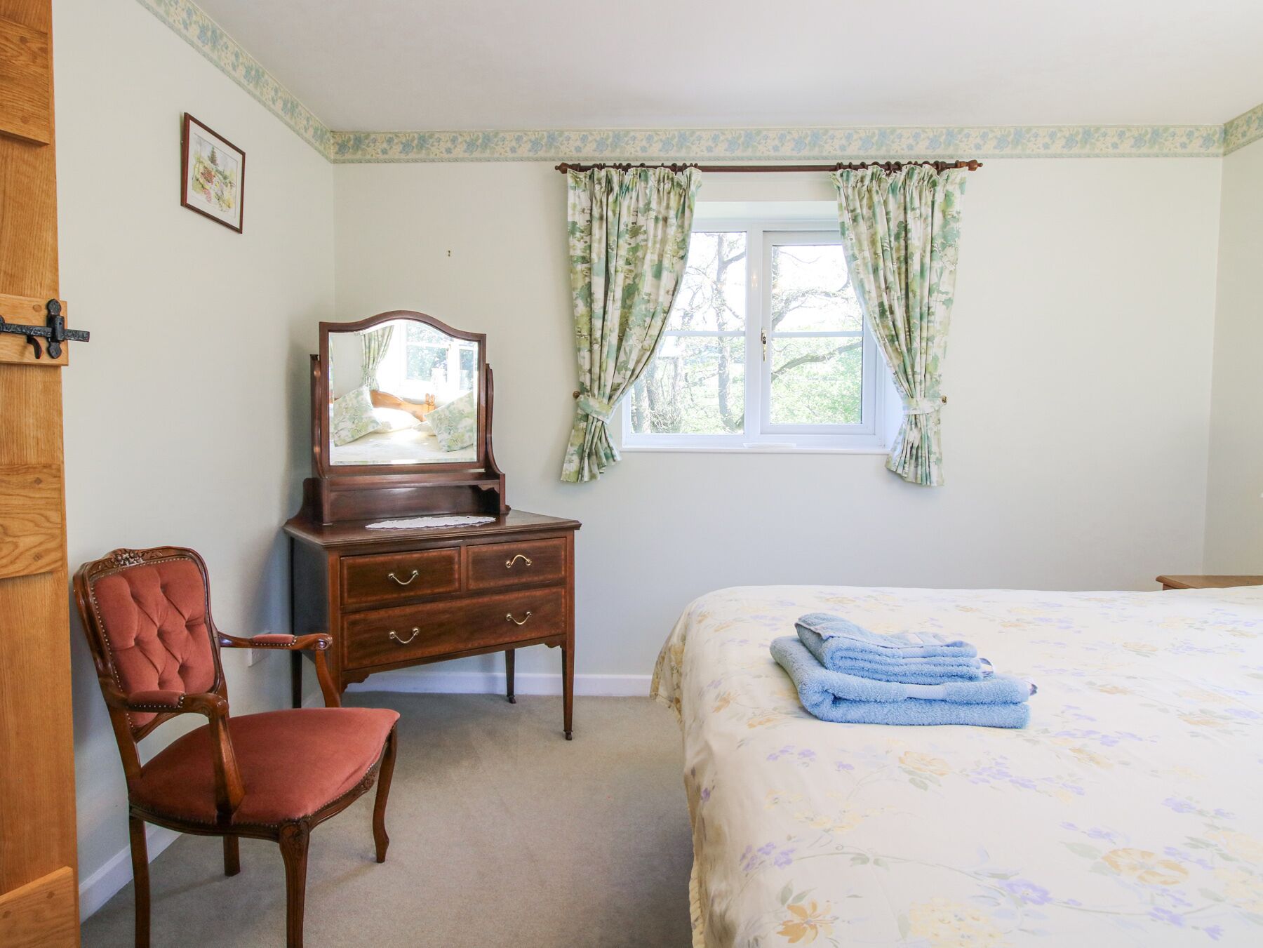 A bedroom with a bed, a wooden dresser with a mirror, a red upholstered chair, and floral curtains.