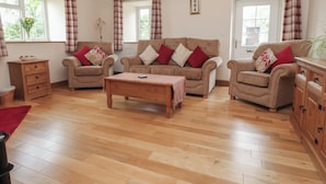 A cozy living room with three sofas, a wooden coffee table, and a cabinet. There are two windows with curtains, a vase with flowers, and a framed picture on the wall. - Bryn Caled (Pwllheli)
