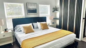 Une chambre moderne avec un grand lit, deux tables de chevet, une petite table, une décoration murale en forme d’étoile et une fenêtre avec des stores.