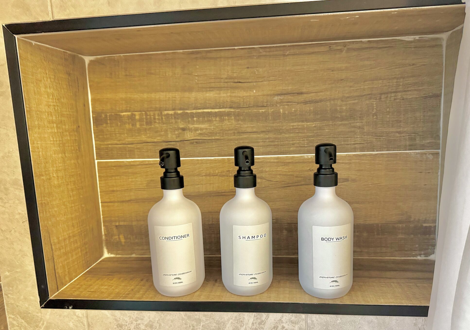 A wooden shelf with three bottles of toiletries labeled conditioner, shampoo, and body wash.