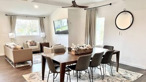 Un salon moderne avec un coin repas, comprenant une grande table en bois, des chaises grises, un ensemble de canapés et un ventilateur de plafond.