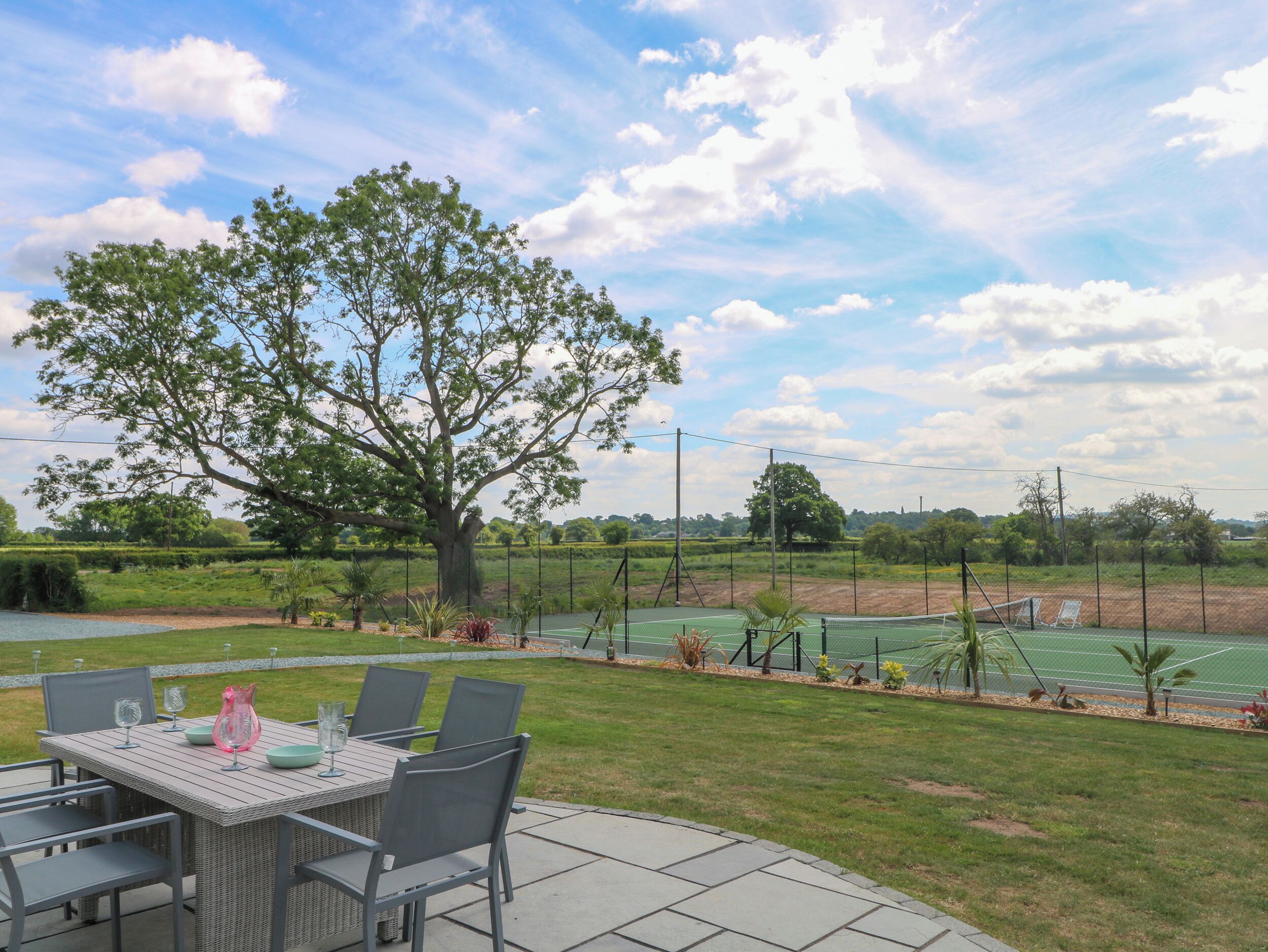 Une terrasse extérieure avec une table et des chaises, entourée d’un court de tennis et d’un grand arbre.