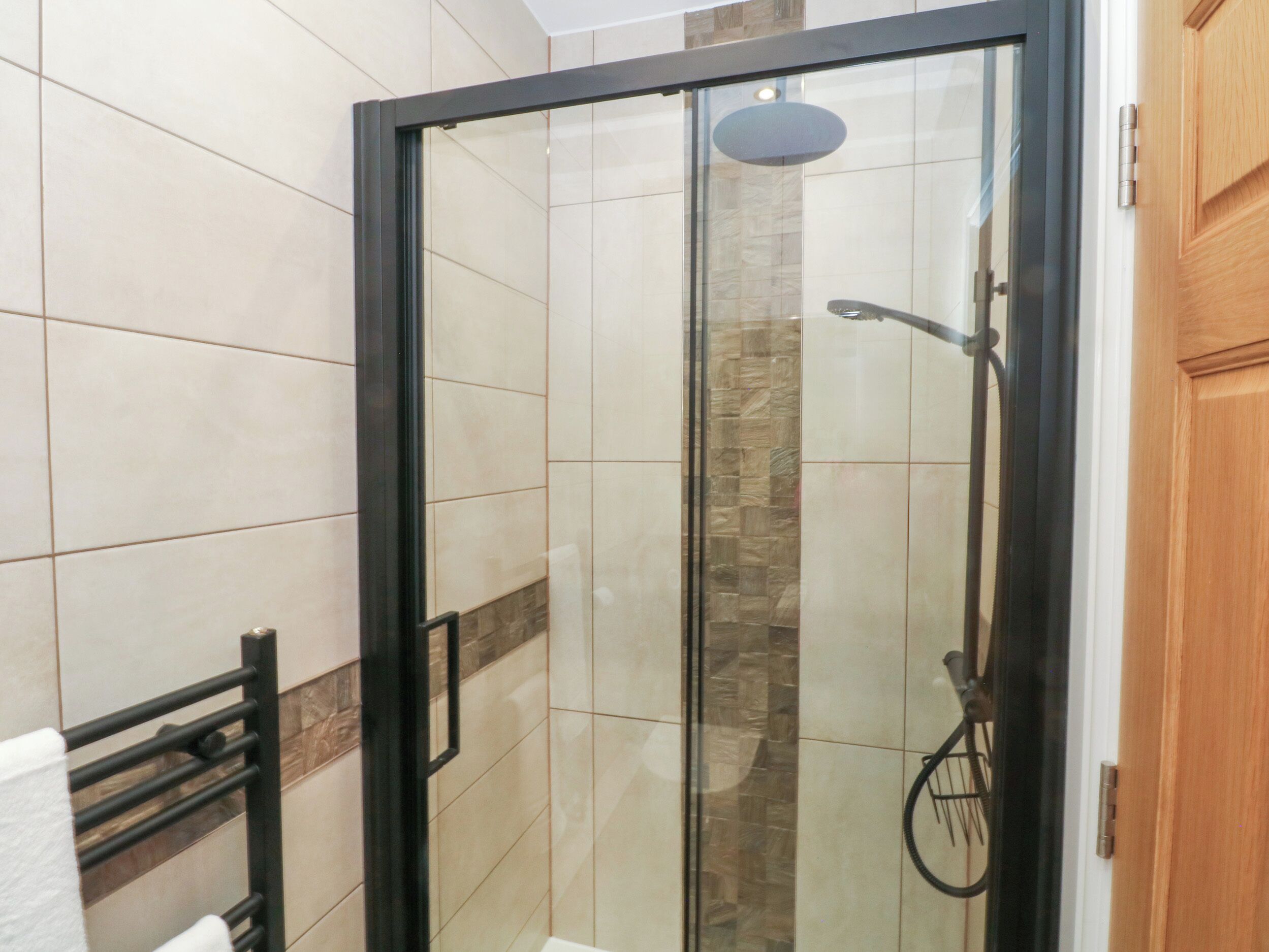 Una ducha con cerramiento de vidrio, un cabezal de ducha tipo lluvia, un perchero y una puerta de madera.