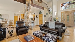 A spacious living room with a stone pillar, leather furniture, a staircase, and a view of the outside. - European Rustic Mountain Retreat in Torreon (Show Low)
