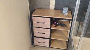A small wooden desk with drawers and a shelf, placed in a corner with a tiled wall and a glass door.